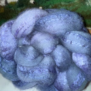 May include: A close-up of a pile of blue and purple wool roving. The wool is soft and fluffy, with variations in color and texture. The background shows a felted landscape with a bunny and sheep.