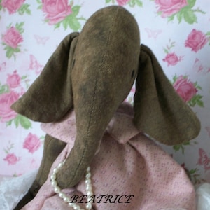 May include: A brown fabric elephant doll wearing a pink dress with a white pearl necklace. The dress has a small white pattern and the text "BEATRICE BLACK CAT & ROSES 2015" is printed on the bottom.