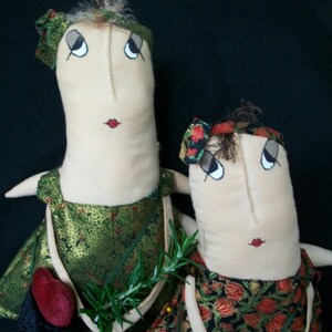 Könnte beinhalten: Zwei handgefertigte Stoffpuppen mit gestickten Gesichtern und floral gemusterten Kleidern. Jede Puppe hält eine kleine Samt Tasche mit einer herzförmigen Klappe. Die Puppen sind vor einem schwarzen Hintergrund platziert.