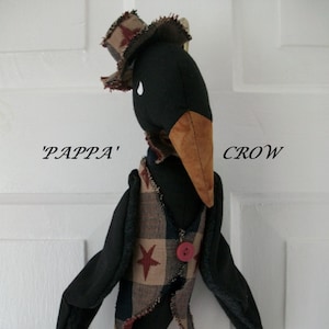Può includere: Una bambola di corvo nero con un becco marrone e un gilet a quadri rosso, bianco e blu. Il corvo indossa un cappello e ha un bottone sul suo gilet. La bambola è etichettata 'Papppa' Crow.