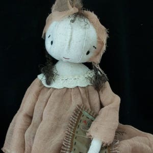 May include: A handmade rag doll with brown hair, wearing a brown dress and holding a small patchwork quilt. The doll has a simple white face with painted eyes and a sweet expression.