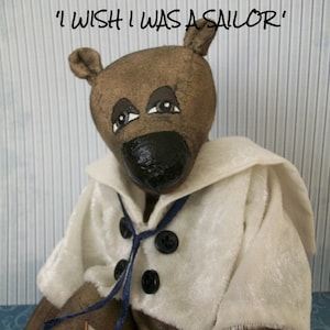 May include: A brown teddy bear wearing a white sailor outfit with a blue ribbon and black buttons. The bear is holding a small wooden sailboat. The text "I wish I was a sailor" is written above the bear.