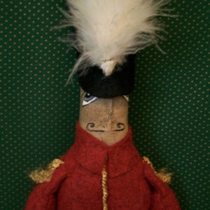 Puede incluir: Un juguete de peluche de un soldado con un sombrero de plumas blancas, un sombrero negro, una chaqueta roja, ribetes dorados y una cara marrón. El juguete está de pie sobre un fondo verde.