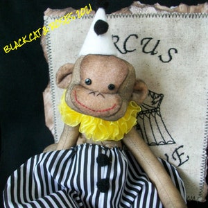 May include: A plush monkey toy wearing a white and black clown hat, a yellow ruffle collar, and black and white striped pants. The toy is sitting on a piece of fabric with the word "Circus" embroidered on it. The text "Cheeky Chimp!" is written below the toy.