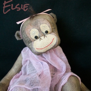 May include: A handmade plush monkey doll named Elsie, wearing a pink tulle dress. The doll has a brown face with painted features and a pink bow in its hair. The doll is sitting on a black background. The text "Black Cat & Roses 2012" is written on the dress.