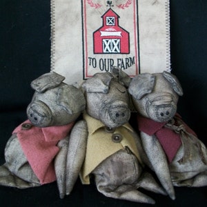 May include: Three gray fabric pig figurines wearing different colored clothing. The pig on the left is wearing a red shirt, the pig in the middle is wearing a yellow shirt, and the pig on the right is wearing a maroon shirt. A sign behind the pigs reads "Welcome to our farm" with a red barn illustration.