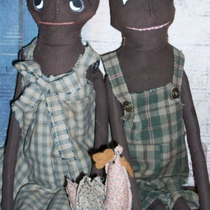 May include: Two handmade rag dolls with brown skin and black eyes. The doll on the left is wearing a plaid dress with a bow. The doll on the right is wearing plaid overalls. Both dolls are holding a small, pink and white fabric chicken.