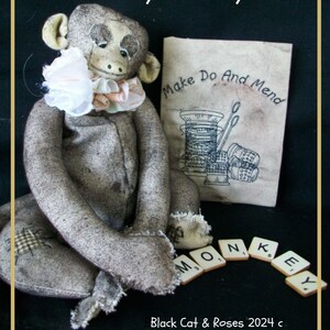 May include: A gray felt monkey toy with a pink and white fabric collar. The monkey is sitting on a black surface with a book titled "Make Do And Mend" and the word "MONKEY" spelled out with letter tiles.