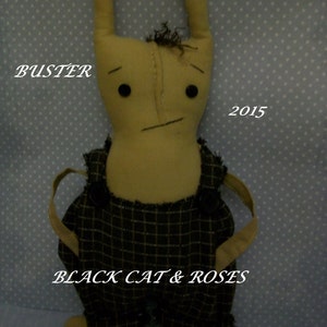 Puede incluir: Una muñeca de trapo hecha a mano con brazos y piernas largos, que lleva un mono con estampado de cuadros. La muñeca tiene una cara cosida con ojos negros y una boca cosida. La muñeca está etiquetada con "Buster" y "Black Cat & Roses" con el año "2015".