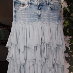 May include: A denim skirt with a light blue wash, featuring a layered, ruffled skirt in a soft, muted blue fabric. The skirt has a classic five-pocket design and a zipper closure.
