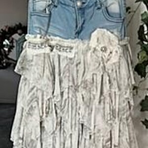 May include: A unique skirt made from repurposed light-wash denim jeans and layered, ruffled fabric. The skirt features the jean's pockets and waistband, with a decorative flower and embellishments. The overall design is a blend of casual and bohemian styles.