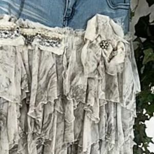 May include: A layered, ruffled skirt with a denim waistband. The skirt features a gray and white marbled pattern and is adorned with lace and a decorative flower with a brooch. The skirt's design is bohemian and romantic.
