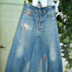 May include: A light blue denim skirt, repurposed from jeans, hangs on a hanger. The skirt features a button fly, pockets, and decorative patches. Faded areas and distressed details add to the vintage aesthetic.