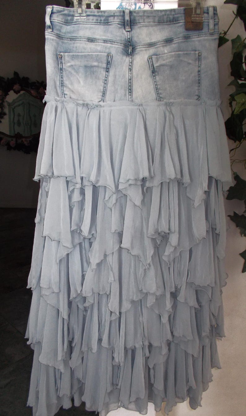 May include: A light blue denim skirt with a layered, ruffled chiffon design. The skirt features a denim waistband with back pockets and a cascading, tiered chiffon skirt. The skirt is hanging on a hanger.