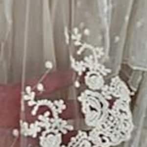 May include: Close-up of a sheer, cream-colored fabric with delicate white floral lace detailing. The lace features intricate rose and leaf patterns along the edge. The fabric has a soft, flowing appearance, suggesting it might be part of a garment or decorative item.