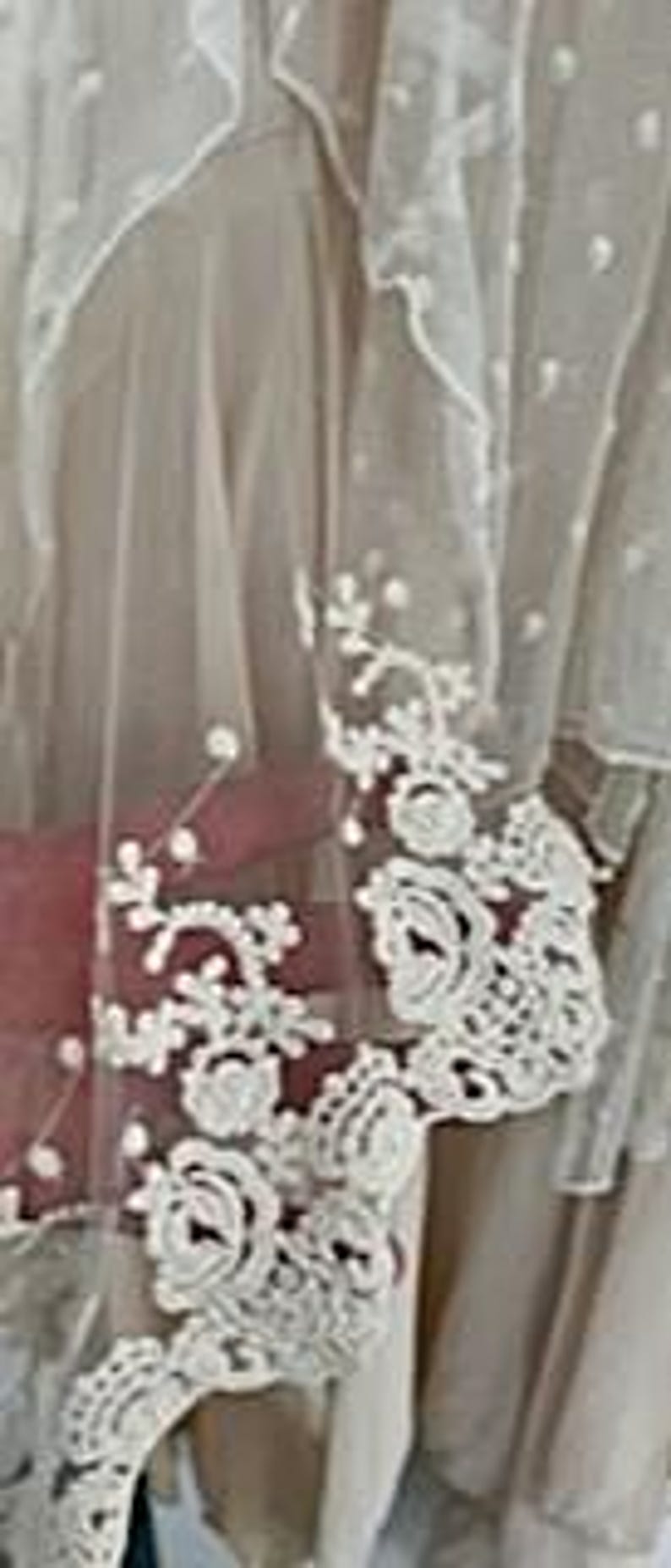 May include: Close-up of a sheer, cream-colored fabric with delicate white floral lace detailing. The lace features intricate rose and leaf patterns along the edge. The fabric has a soft, flowing appearance, suggesting it might be part of a garment or decorative item.