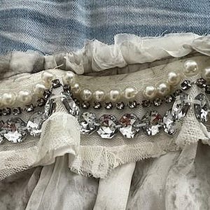 May include: Close-up of a decorative textile with layers of white fabric, pearls, and sparkling rhinestones. The design includes a denim blue fabric at the top, with a layered, ruffled effect. The embellishments are arranged in horizontal rows.
