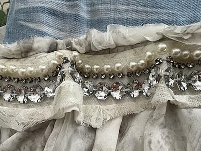 May include: Close-up of a decorative textile with layers of white fabric, pearls, and sparkling rhinestones. The design includes a denim blue fabric at the top, with a layered, ruffled effect. The embellishments are arranged in horizontal rows.