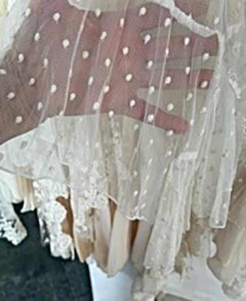 May include: Close-up of a sheer, off-white fabric with white polka dots. The fabric is layered over other white and cream-colored materials, creating a textured, layered effect. The image shows a glimpse of a hand behind the fabric.