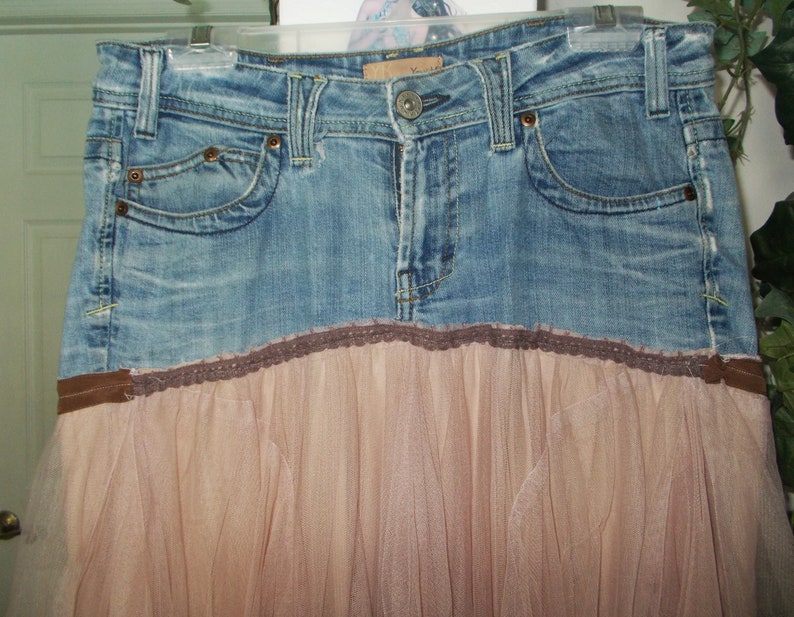 May include: A denim skirt with a brown lace trim and a tulle overlay. The skirt is a light blue color and has a brown belt.