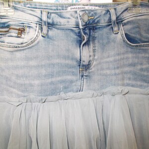 May include: Light blue denim skirt with a zipper pocket and a light blue tulle ruffle. The skirt has a button closure and belt loops. The brand name "ZARA BASIC" is visible on the waistband.