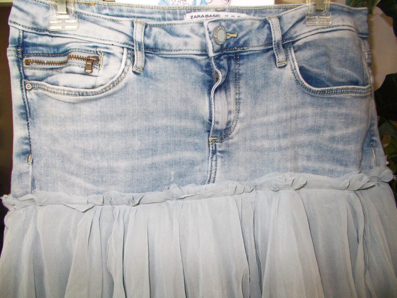May include: Light blue denim skirt with a zipper pocket and a light blue tulle ruffle. The skirt has a button closure and belt loops. The brand name "ZARA BASIC" is visible on the waistband.
