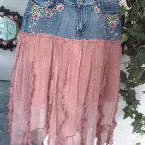 Op de afbeelding: Een spijkerbroekrok met een roze, gelaagde chiffon rok. De spijkerbroek tailleband heeft geborduurde bloemmotieven met kleurrijke kralen. De rok hangt aan een witte hanger.