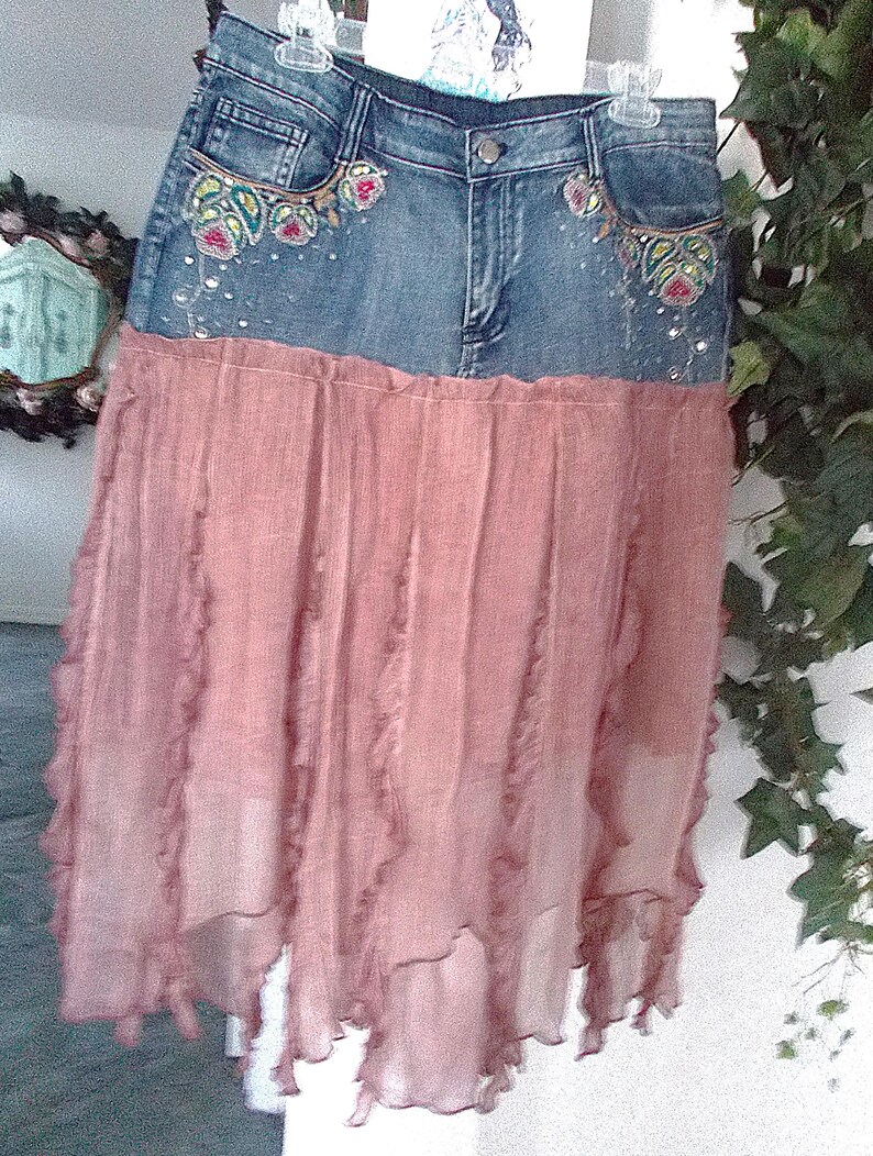 Op de afbeelding: Een spijkerbroekrok met een roze, gelaagde chiffon rok. De spijkerbroek tailleband heeft geborduurde bloemmotieven met kleurrijke kralen. De rok hangt aan een witte hanger.