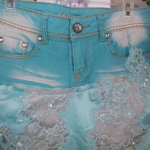 May include: Light blue denim skirt with a satin bottom and lace detailing. The skirt features a button closure, rhinestone accents on the pockets, and a faded wash effect. The lace is embellished with sequins.