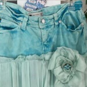 Puede incluir: Una falda vaquera turquesa con un efecto tie-dye azul claro. La falda presenta un dobladillo de gasa verde claro con volantes y una flor de tela decorativa con un centro de cuentas. La falda est&aacute; colgada en una percha.