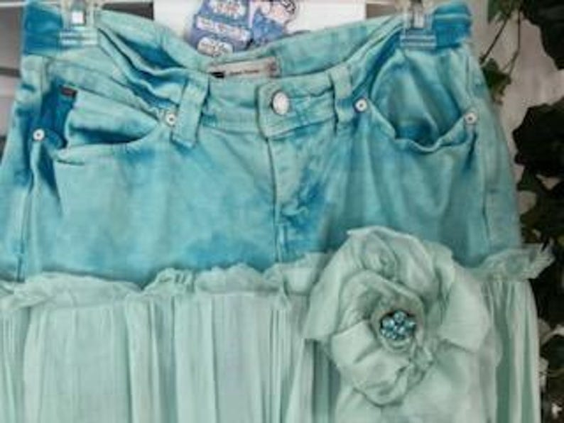 Puede incluir: Una falda vaquera turquesa con un efecto tie-dye azul claro. La falda presenta un dobladillo de gasa verde claro con volantes y una flor de tela decorativa con un centro de cuentas. La falda est&aacute; colgada en una percha.