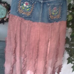 Op de afbeelding: Een spijkerbroekrok met een roze, vloeiende, gelaagde onderkant. De rok heeft twee achterzakken met geborduurde bloemmotieven. De denim is een medium wassing en de roze stof heeft een gerimpelde rand.