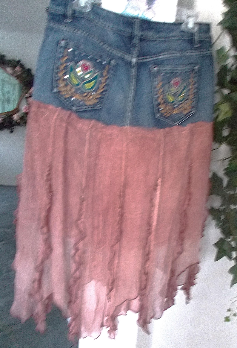 Op de afbeelding: Een spijkerbroekrok met een roze, vloeiende, gelaagde onderkant. De rok heeft twee achterzakken met geborduurde bloemmotieven. De denim is een medium wassing en de roze stof heeft een gerimpelde rand.