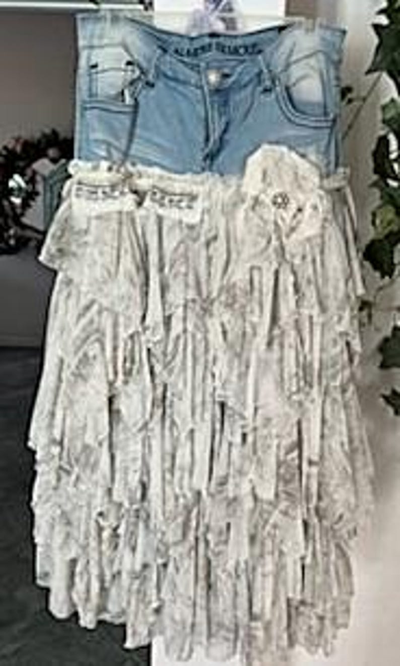 May include: A unique skirt made from repurposed light-wash denim jeans and layered, ruffled fabric. The skirt features the jean's pockets and waistband, with a decorative flower and embellishments. The overall design is a blend of casual and bohemian styles.