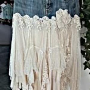 May include: A denim skirt with a layered, off-white lace and fabric design. The skirt features a classic blue jean waistband and pockets, transitioning into a cascading, bohemian-style lace skirt. The lace has intricate floral patterns.