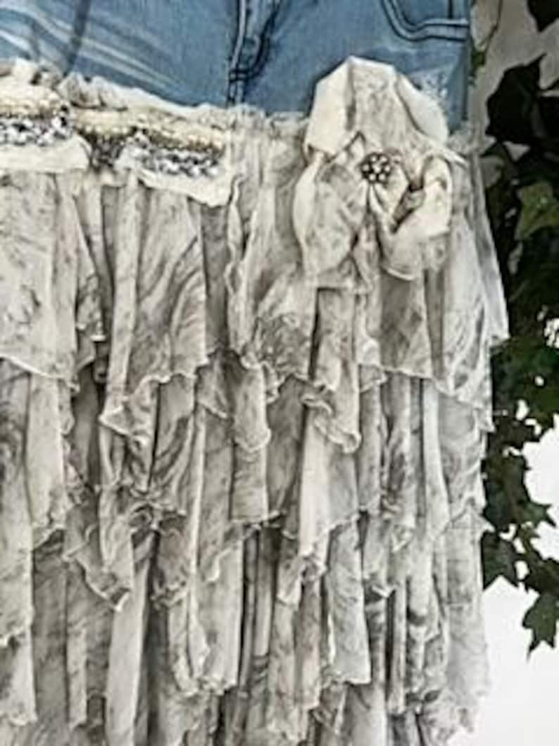 May include: A layered, ruffled skirt with a denim waistband. The skirt features a gray and white marbled pattern and is adorned with lace and a decorative flower with a brooch. The skirt's design is bohemian and romantic.