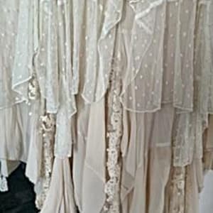 May include: Close-up of a cream-colored, layered skirt with multiple tiers of fabric. The skirt features ruffles, lace, and a dotted pattern, creating a flowing, textured look. The skirt appears to be made of lightweight material.