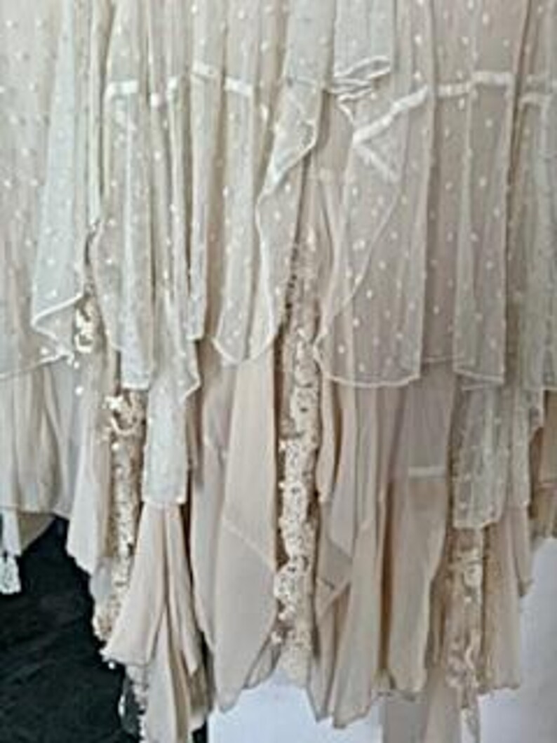 May include: Close-up of a cream-colored, layered skirt with multiple tiers of fabric. The skirt features ruffles, lace, and a dotted pattern, creating a flowing, textured look. The skirt appears to be made of lightweight material.