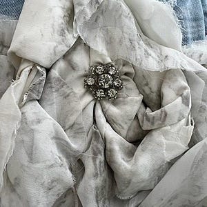 May include: Close-up of a fabric flower with a rhinestone center. The flower is made of layered, ruffled fabric in shades of white and gray, with a marbled pattern. The background includes denim.