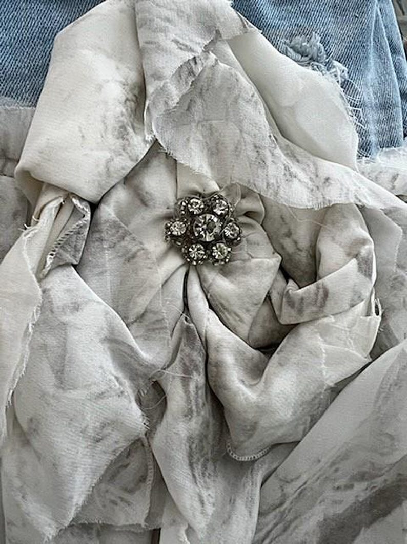 May include: Close-up of a fabric flower with a rhinestone center. The flower is made of layered, ruffled fabric in shades of white and gray, with a marbled pattern. The background includes denim.