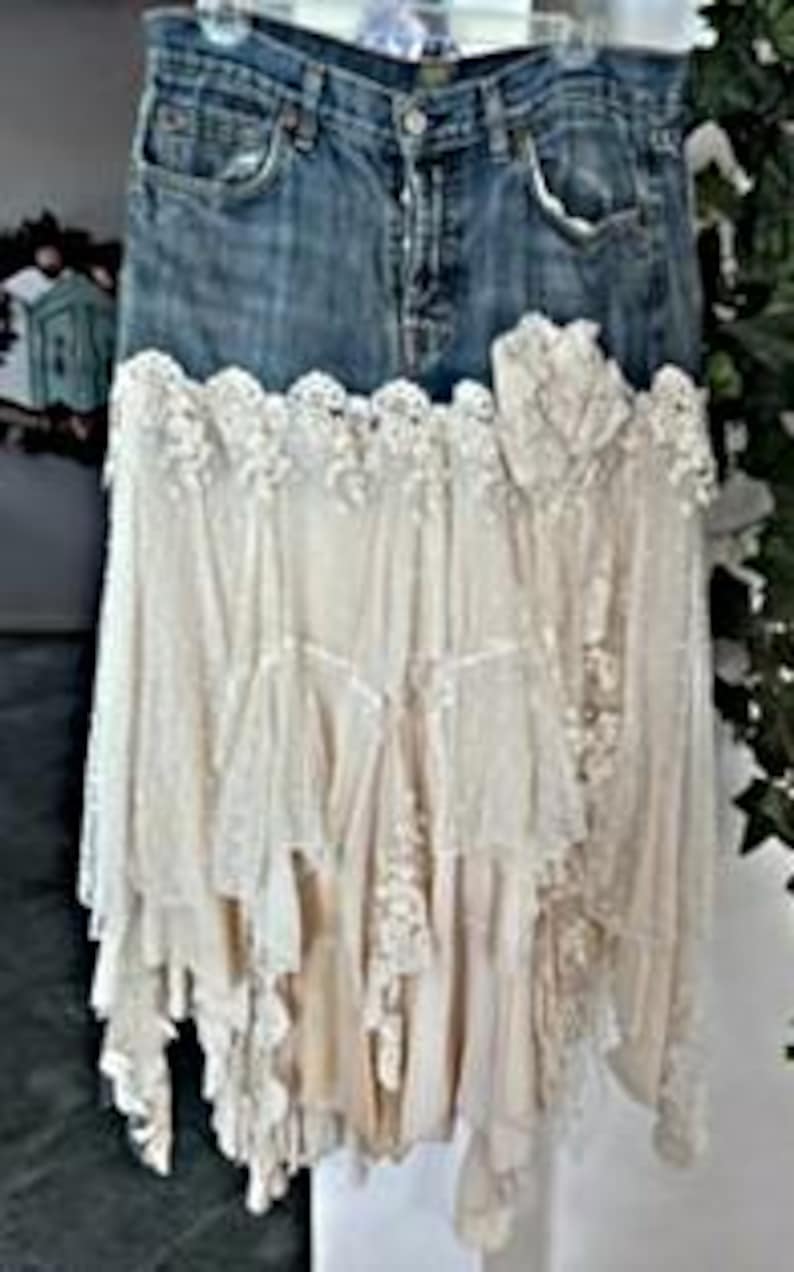 May include: A denim skirt with a layered, off-white lace and fabric design. The skirt features a faded blue denim waistband and a cascading, asymmetrical lace overlay. The lace has intricate floral patterns and a vintage aesthetic.