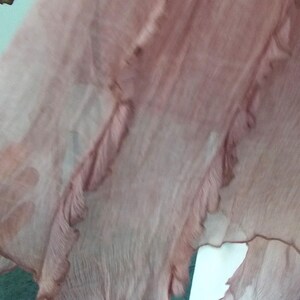 Op de afbeelding: Een vloeiende, transparante stof in tinten van stoffig roze. De stof heeft een gelaagd ontwerp met gerimpelde randen, waardoor een delicate en luchtige uitstraling ontstaat. Het materiaal lijkt lichtgewicht en mogelijk zijde of een vergelijkbare vezel.