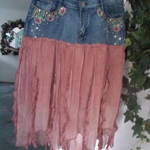 Op de afbeelding: Een spijkerbroekrok met een vervaagde blauwe wassing en een vloeiende, gelaagde, stoffige roze rok. De jeans tailleband heeft geborduurde bloemmotieven met pailletten. De rok hangt aan een hanger.