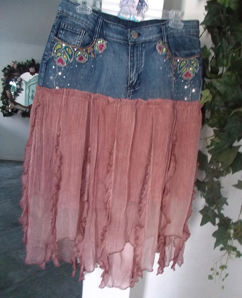 Op de afbeelding: Een spijkerbroekrok met een vervaagde blauwe wassing en een vloeiende, gelaagde, stoffige roze rok. De jeans tailleband heeft geborduurde bloemmotieven met pailletten. De rok hangt aan een hanger.