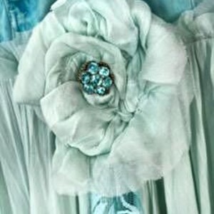Puede incluir: Una gran flor de tela azul claro con m&uacute;ltiples capas de p&eacute;talos. El centro de la flor presenta un grupo de peque&ntilde;as cuentas redondas de color turquesa. La flor est&aacute; sujeta a una prenda azul claro.