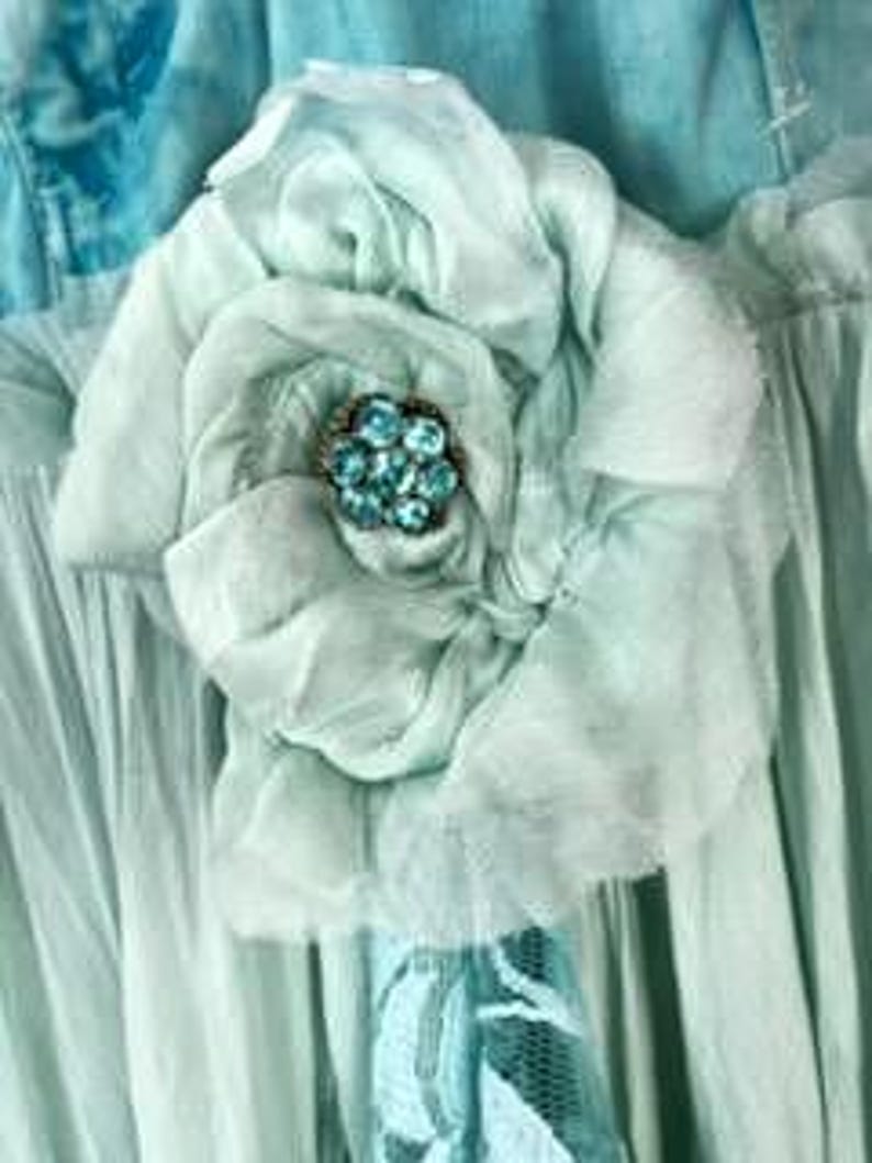 Puede incluir: Una gran flor de tela azul claro con m&uacute;ltiples capas de p&eacute;talos. El centro de la flor presenta un grupo de peque&ntilde;as cuentas redondas de color turquesa. La flor est&aacute; sujeta a una prenda azul claro.