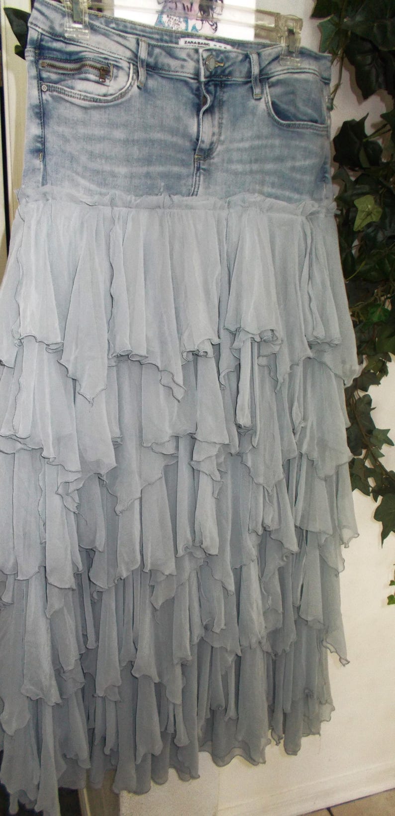 May include: A denim skirt with a light wash, paired with a layered, flowing, gray chiffon skirt. The skirt has a zipper and button closure. The chiffon skirt has multiple layers of ruffles.