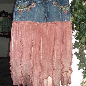 Op de afbeelding: Een spijkerbroekrok met een roze, geplooid chiffon onderstuk. De spijkerbroektop heeft geborduurde bloemmotieven op de zakken en verspreide versieringen. De rok hangt aan een doorzichtige hanger.