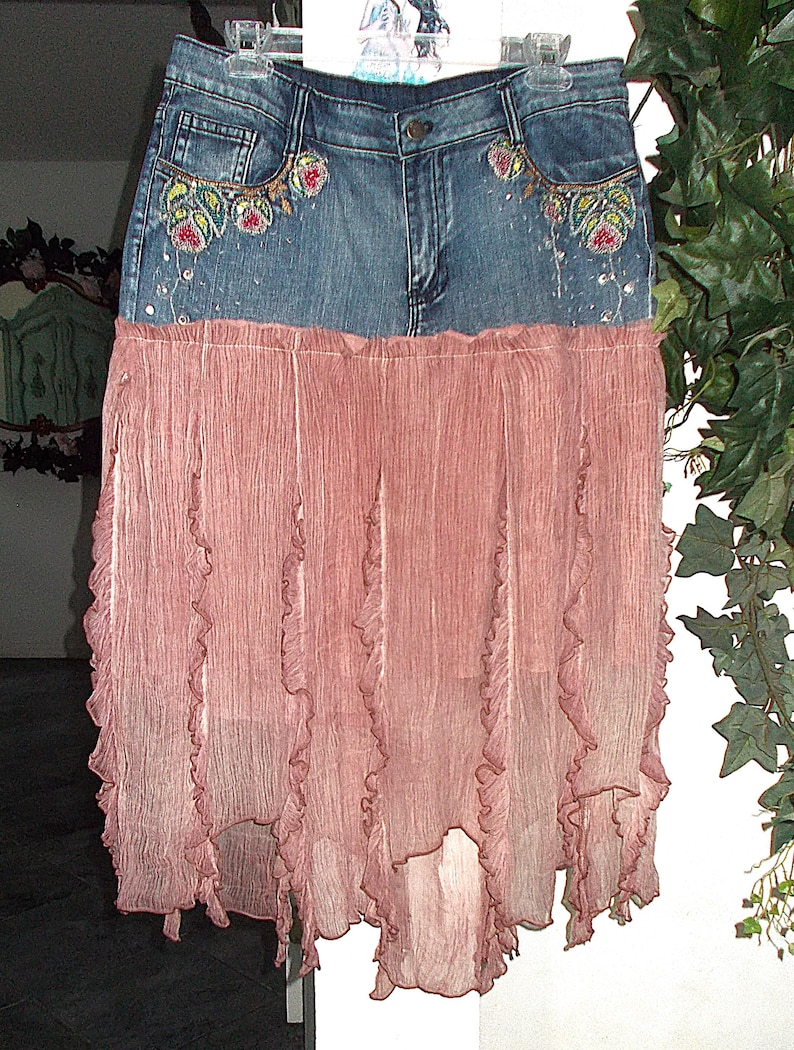Op de afbeelding: Een spijkerbroekrok met een roze, geplooid chiffon onderstuk. De spijkerbroektop heeft geborduurde bloemmotieven op de zakken en verspreide versieringen. De rok hangt aan een doorzichtige hanger.