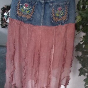 Op de afbeelding: Een spijkerbroekrok met een vervaagde blauwe wassing en een roze, vloeiende stoffen onderkant. Het denimgedeelte heeft geborduurde bloemmotieven op de achterzakken. De rok heeft een uniek, gelaagd ontwerp dat casual denim combineert met een zacht, vloeiend materiaal.